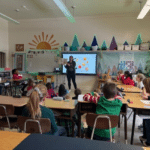 A teacher stands in front of her 2nd grade class. Behind her is a diagram showing different types of winter squash.