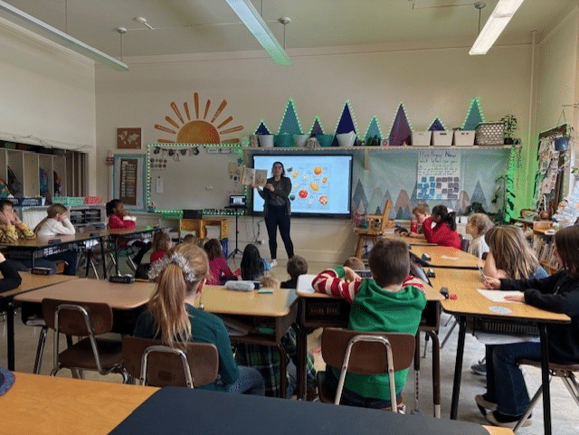 A teacher stands in front of her 2nd grade class. Behind her is a diagram showing different types of winter squash.