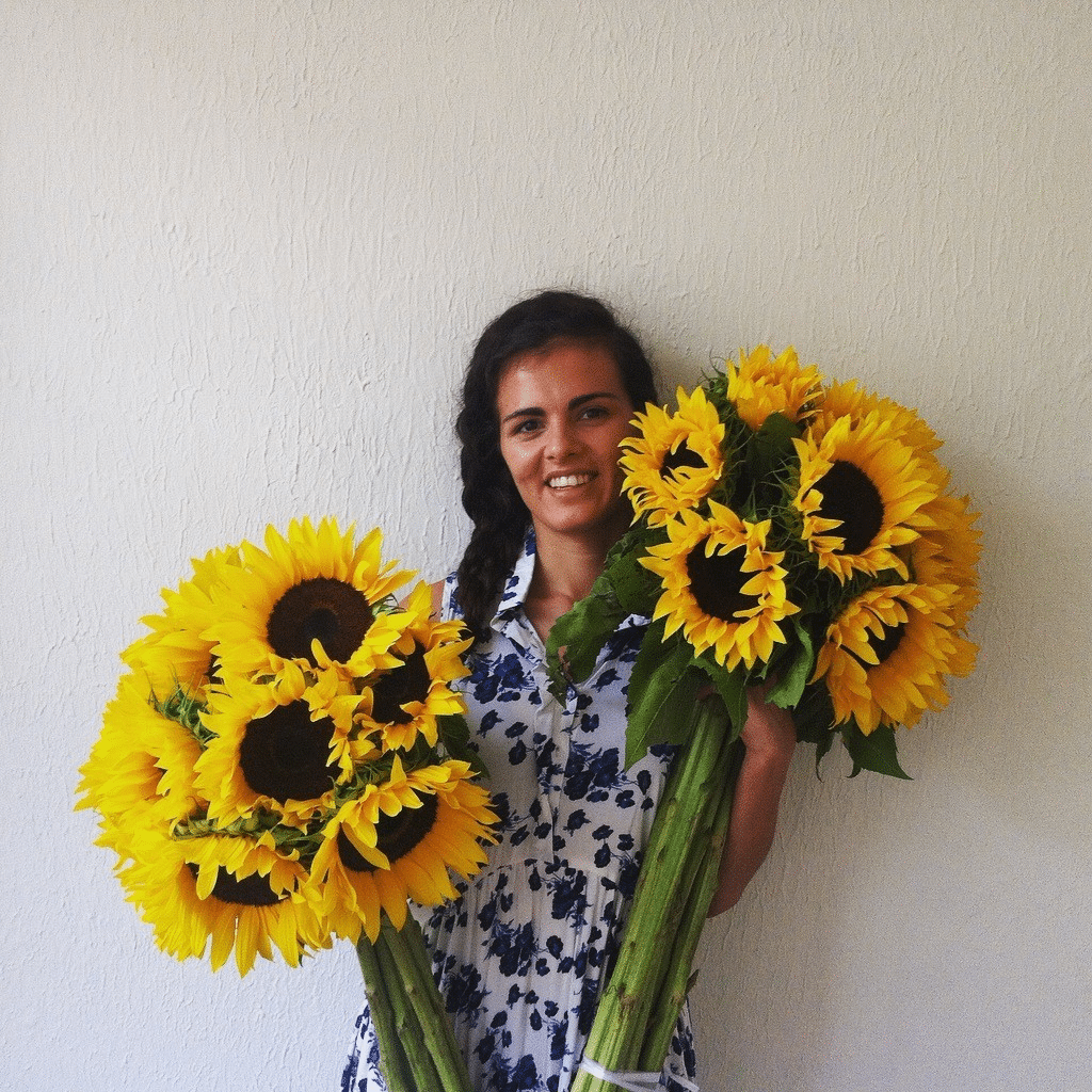 Ines Chapela stands against a wall, smiling and holding two enormous bouquets of yellow sunflowers.