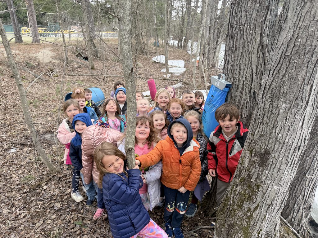 Children smiling outdoors near a tree with sap collection equipment