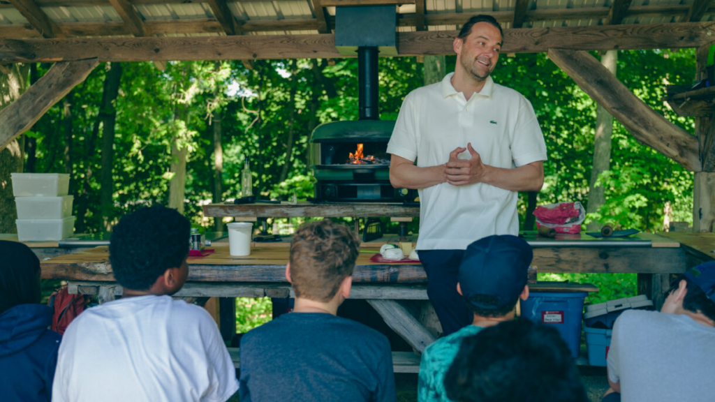 Man teaching outdoors with wood-fired oven and students