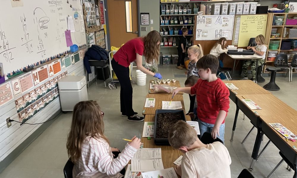 The photo depicts children sitting at their school table. Ellie Hoffman, local food educater through cornell cooperative extension of essex county, is shown planting seeds in cups.